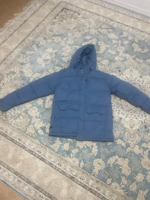 Blå dunjacka från Tuxer med huva - Säljer en blå dunjacka från Tuxer i storlek M. Jackan har huva med snörning, stora fickor framtill och Tuxer-logga på ärmen. Perfekt för kalla dagar och riktigt skön att bära. Stängs med både dragkedja och knappar.