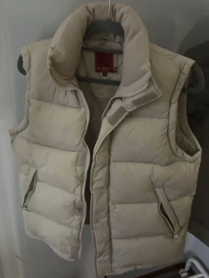 Beige dunväst från Blend - Snygg beige dunväst från Blend med hög krage och dragkedja framtill. Västen har två fickor med dragkedja och är quiltad för extra värme. Perfekt att slänga över en hoodie när det är kyligt ute.