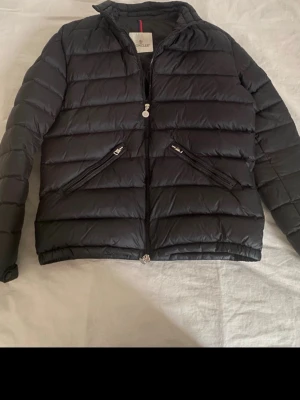 Svart Moncler pufferjacka - Säljer en svart dun jacka från Moncler med klassisk quiltad design och två snedställda fickor med dragkedja. Jackan har Moncler-logga på ärmen och hel dragkedja framtill. Perfekt för kyliga dagar och riktigt snygg till streetwear.