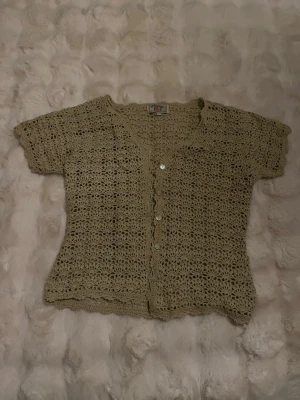 Beige virkad kofta från Soris - Virkad beige kofta från Soris med korta ärmar och v-ringning. Koftan har dekorativa knappar framtill och ett luftigt, öppet mönster som ger en bohemisk vibe. Perfekt att slänga över ett linne under varma dagar.
