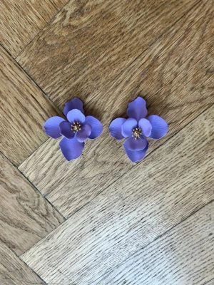Lila blommiga örhängen i plast - Säljer ett par stora lila örhängen formade som blommor med gulddetaljer i mitten. Örhängena är ca 4,5 cm i diameter och har en matt finish. Perfekta för att ge din outfit en färgklick och lite flower power-vibe.