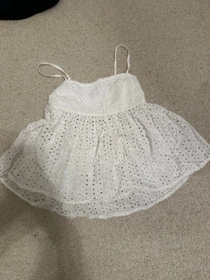 Vit broderad linnetopp med spets - Söt vit linnetopp med tunna axelband och broderade hålmönster över hela tyget. Toppen har en luftig och flowy passform med volangkant nedtill och fina spetsdetaljer längs överkanten. Perfekt för varma dagar och sommarvibbar.