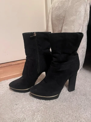 Svarta stövletter med klack och gulddetalj - Svarta stövletter i mocka med hög klack och spetsig tå. Skorna har dragkedja på sidan och en snygg guldfärgad rand som detalj vid tån. Stilrena och eleganta! Mycket bra skick, storlek 37. Säljs på flera sidor, först till kvarn. 