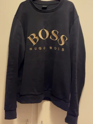 Svart sweatshirt från Hugo Boss - Säljer en svart sweatshirt från Hugo Boss med stor guldfärgad BOSS-logga broderad på bröstet. Tröjan har rund halsringning och långa ärmar. Perfekt för dig som vill ha en stilren och trendig look.