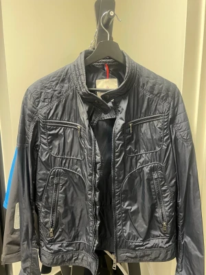 Svart vindjacka från Moncler - Snygg svart vindjacka från Moncler med flera dragkedjefickor framtill. Jackan har en sportig look med quiltade detaljer och Moncler-logga på ärmen. Tillverkad i 100% nylon som ger en lätt och smidig känsla. Perfekt för dig som gillar stilren och funktionell design.