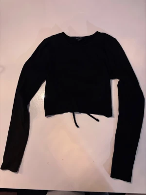 Svart croppad topp från Monki - Säljer en svart croppad topp från Monki i ribbad bomullsmix. Toppen har långa ärmar, rund halsringning och öppen rygg med knytband bak. Perfekt för dig som gillar trendiga detaljer och enkel stil.