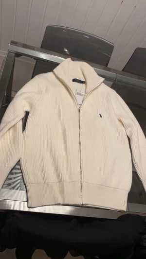 Vit stickad kofta från Polo Ralph Lauren - Säljer en clean vit stickad kofta från Polo Ralph Lauren med dragkedja framtill och hög krage. Klassisk ribbad struktur och diskret logga broderad på bröstet. Perfekt till lager på lager under höst och vinter.