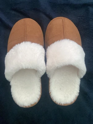 Bruna tofflor med vit fuskpäls - Mysiga bruna tofflor med fluffig vit fuskpäls på insidan och kanten. Perfekta för kalla dagar hemma. Yttermaterialet är mjukt och sulan är i syntet med bra grepp. Passar dig som vill ha både stil och komfort.