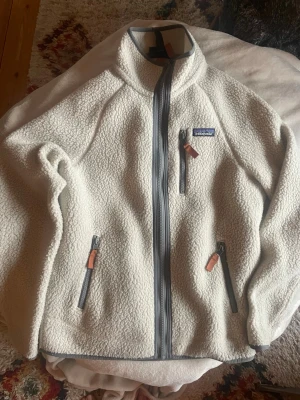 Vit jacka från Patagonia XS - Säljer en vit teddyjacka från Patagonia med grå dragkedja. Använd endast några månader då jag växt ur den. Sitter med normal passform och går ner till midjan. Skick 9/10. Inga fel eller märken. Skicka dm vid frågor.