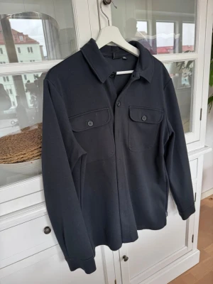 Svart overshirt från Only & Sons - Säljer en svart overshirt från Only & Sons i storlek M. Jackan har två bröstfickor med knappar, klassisk krage och stängs med knappar framtill. Materialet är mjukt och känns som bomull, perfekt för lager på lager och en clean look.