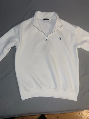 Vit half zip från Polo Ralph Lauren - Säljer en clean vit tröja från Polo Ralph Lauren med half zip och klassisk broderad logga på bröstet. Tröjan har lång ärm och hög krage, perfekt för lager på lager. Skön och stilren, passar till allt.