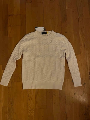 Vit kabelstickad tröja Polo Ralph Lauren - Kabelstickad vit tröja från Polo Ralph Lauren med klassisk rund halsringning och broderad logga på bröstet. Långärmad modell i mjuk bomull som ger en clean och tidlös vibe. Perfekt till jeans eller kjol för en preppy look.