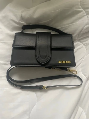 Svart axelväska från Jacquemus - Säljer en stilren svart axelväska från Jacquemus i slätt skinn. Väskan har ett elegant lock med bred rem och guldfärgade metalldetaljer. Justerbar axelrem medföljer och loggan syns i guld på framsidan. Perfekt för dig som gillar minimalistisk design.