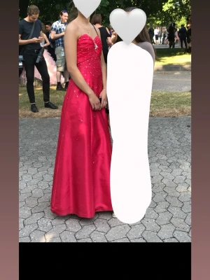 Röd långklänning med strassdetaljer - Säljer en röd långklänning med glittriga strassdetaljer och v-ringad överdel. Klänningen har axelband och en elegant, utsvängd kjol. Perfekt för bal eller student! Materialet är glansigt och ger en lyxig känsla.