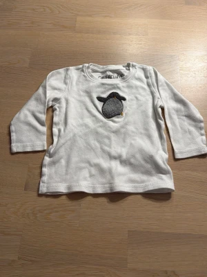 Vit långärmad tröja med pingvin från Next - Söt vit långärmad tröja från Next i 100% bomull, dekorerad med en broderad pingvin på bröstet. Perfekt för små barn som gillar djurmotiv. Mjuk och bekväm passform med rund hals.