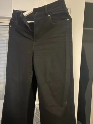 Svarta high waist jeans med wide fit - Säljer ett par svarta jeans med hög midja och breda ben. Klassisk femficksmodell med kopparfärgade nitar och bälteshällor. Materialet är stadigt jeans-tyg som ger en snygg siluett. Perfekta till sneakers eller boots.