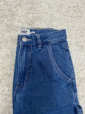 Blå raka jeans från 157 - Säljer ett par klassiska blå jeans från 157 med raka ben och hög midja. Jeansen har fem fickor, bälteshällor och stängs med knapp och dragkedja. Materialet är slitstark denim i bomull och passformen är normal. Perfekta till en avslappnad look.