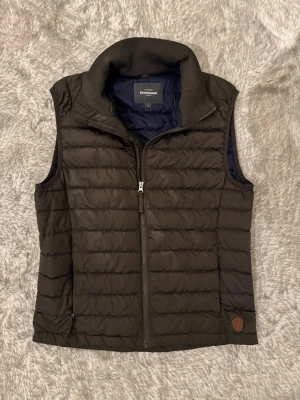 Brun dunväst - Brun dunväst från Dressmann i storlek S med quiltad design och dragkedja framtill. Västen har hög krage och två sidofickor med dragkedja. 