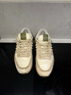 Valentino Rockrunner beige sneakers - Säljer ett par beige Valentino Rockrunner sneakers med ikoniska nitar på sidorna och hälen. Skorna har snörning, rund tå och en mix av mocka och skinn i ovandelen. Snygg kontrast mellan olika beige toner och coola detaljer som ger en lyxig vibe. Den vänstra skorn har Tyvär tappat en nit, det är också små hål i skorna på hälarna ”därför så lågt pris” men just nu sitter det små laglappar som ska sitta där och det känns inget när man går. Pris ej hugget i sten!! SKRIV VID MINSTA FUNDERING!! 