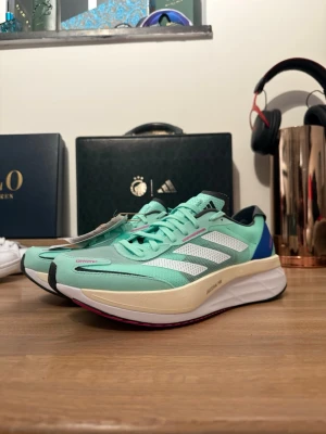 Mintgröna Adidas Adizero sneakers - Säljer ett par Adidas Adizero sneakers i mintgrönt med vita och svarta detaljer samt blå och rosa inslag. Skorna har mesh-ovandel för bra ventilation, snörning och en chunky, vit sula med rosa och svart yttersula. Perfekta för dig som gillar sportig stil och vill sticka ut. INTE fejk, morsan jobbar där, vi vill bara bli av med skor