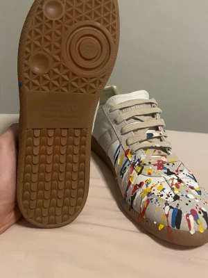 Maison Margiela sneakers med färgstänk - Maison Margiela sneakers i vitt och beige skinn och mocka med unikt färgstänkt mönster i rött, blått, gult, svart och vitt. Skorna har klassisk gummisula, snörning och en stilren, rund tå. Perfekta för dig som vill sticka ut med en kreativ vibe.