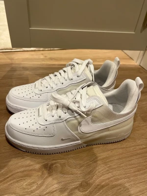 Nike Air Force 1 vita sneakers - Säljer ett par fräscha Nike Air Force 1 sneakers i helvitt. Skorna har klassisk perforerad tå, snörning och en mix av skinn och syntetmaterial med genomskinliga detaljer på sidan och hälen. Snygg och stilren modell som passar till allt.