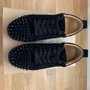 Svarta sneakers från Christian Louboutin - Säljer ett par svarta sneakers från Christian Louboutin i sammet med coola svarta nitar på tån och klassisk röd sula. Skorna har snörning och insida i beige skinn. Perfekta för dig som vill sticka ut med exklusiv design och edgy detaljer.