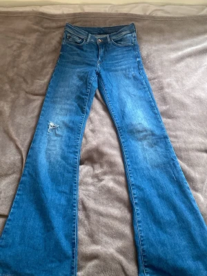Blå flare jeans från H&M - Säljer ett par blåa flare jeans från H&M med hög midja och klassisk femficksdesign. Jeansen har en snygg bootcut passform och är tillverkade i bomull med lite stretch. Det finns en sliten detalj på ena benet som ger en cool touch. Det går att spretta upp byxbenet så jeansen blir längre. 