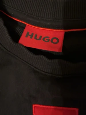 Svart sweatshirt från HUGO - Svart sweatshirt från HUGO med röd logotyp-patch på bröstet. Tröjan har rund halsringning, ribbade muddar och är långärmad. Perfekt för en clean och stilren look. Materialet är mjuk bomull som känns skönt mot huden haft den i 3 månader storlek XS sitter bra 