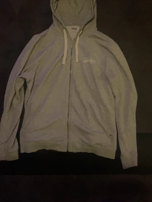 Grå zip-hoodie – stilren & bekväm - Snygg grå hoodie med dragkedja, perfekt för både vardag och chill. Klassisk och tidlös design med diskret broderad logga på bröstet som ger ett rent och stilfullt intryck.  ✔ Mjuk och skön kvalitet ✔ Hel dragkedja – lätt att matcha och använda året runt ✔ Neutral grå färg som passar till allt ✔ Huva med snören för extra komfort  