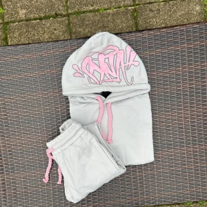 Syna World grå och rosa tracksuit. - Grå hoodie från SNA med stor rosa graffiti-logga på huvan och matchande rosa snören. Tröjan har en klassisk loose passform och är tillverkad i mjuk bomull, perfekt för chill dagar. Snygg streetwear-vibe med unika detaljer som sticker ut.