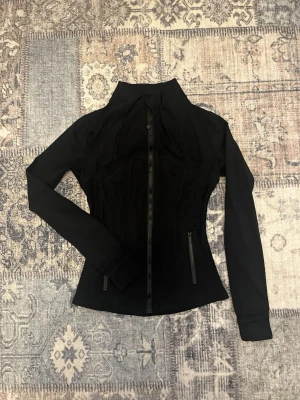 Define Jacket - Snygg och figurnära Define Jacket från Lululemon i klassisk svart färg. Tillverkad i stretchigt, mjukt och följsamt material som ger en smickrande passform och hög komfort. Perfekt både för träning och till vardags.  Jackan har: 	•	Hellång dragkedja 	•	Hög krage 	•	Praktiska fickor med dragkedja 	•	Formande sömmar som ger en stilren siluett  I mycket fint skick, sparsamt använd. Inga hål, fläckar eller noppor.  Ett måste i garderoben för dig som gillar stilrena träningsplagg av hög kvalitet.