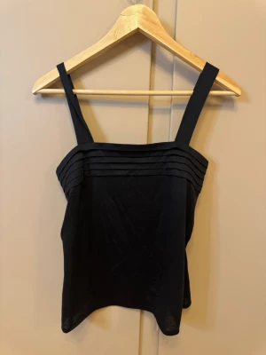 Svart linnetopp med breda axelband - En stilren svart linnetopp med breda axelband och dekorativa horisontella veck framtill. Toppen har rak passform och är tillverkad i ett mjukt tyg som känns skönt mot huden. Perfekt att matcha med jeans eller kjol för en enkel och snygg look.