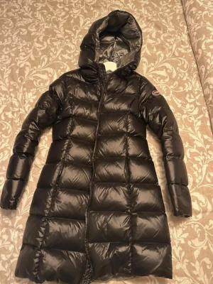 Svart lång pufferjacka från Colmar - Säljer en svart lång pufferjacka från Colmar med huva och dragkedja. Jackan har en glansig finish och breda quiltade paneler som ger en riktigt snygg look. Perfekt för kalla dagar och riktigt skön att bära. Klassisk Colmar-logga på ärmen.