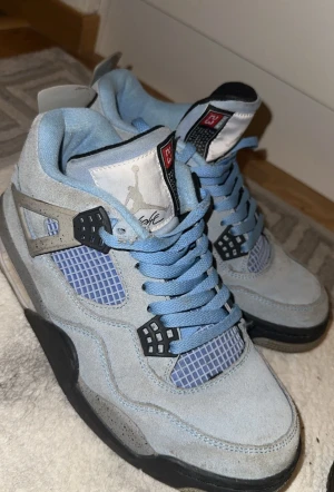 Nike Air Jordan 4 University Blue - Säljer ett par Nike Air Jordan 4 University Blue sneakers i ljusblå mocka med svarta och grå detaljer. Skorna har meshpaneler, snörning och den ikoniska Jumpman-loggan på hälen. Snygg kontrast mellan den blå färgen och de stänkta grå partierna.