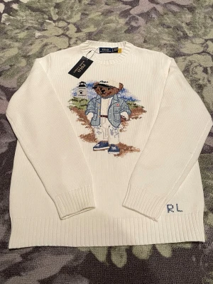 Vit stickad tröja Polo Ralph Lauren - Säljer en vit stickad tröja från Polo Ralph Lauren med ikonisk björn på bröstet. Tröjan har rund hals, ribbade muddar och RL-broderi vid nederkanten. Tillverkad i mjuk bomull och perfekt för dig som vill ha en klassisk men lekfull look.