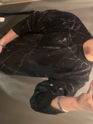 Svart mönstrad sweatshirt från Gymshark - Säljer en svart sweatshirt från Gymshark med grått marmorerat mönster. Tröjan har rund halsringning och långa ärmar. Perfekt för dig som gillar en sportig och avslappnad stil.