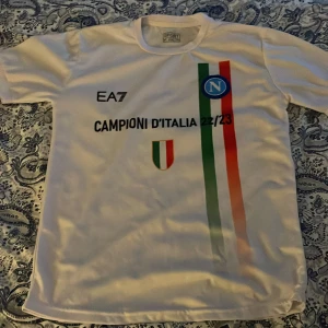 Vit Napoli t-shirt EA7 XXL - Säljer en vit t-shirt från EA7 i storlek M/L med tryck 'Campioni d'Italia 22/23' och Napoli-logga. T-shirten har korta ärmar och färgglada ränder i grönt, vitt och rött på framsidan, samt ett italienskt mästerskapsmärke. Perfekt för fotbollsfans! Tröjan sitter ganska så tajt så den sitter mer som en S/M