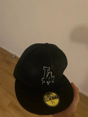 Svart LA New Era 59Fifty keps - Säljer en svart New Era 59Fifty keps med LA-logga i vitt framtill. Klassisk modell med böjd skärm och broderade detaljer på sidorna och bak. Märkt med Cooperstown Collection och har en stilren look som passar streetwear. Perfekt för dig som gillar sportiga accessoarer.