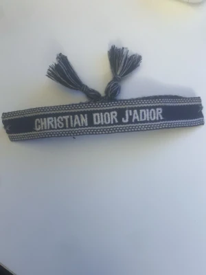 Christian Dior J'Adior armband - Snyggt armband från Christian Dior med texten 'J'Adior' i vitt på en mörkblå bakgrund. Armbandet har två tofsar och är vävt i ett mjukt material som känns lyxigt och trendigt. Perfekt accessoar för att lyfta din outfit.