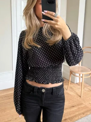 Svart prickig blus - Säljer en svart croppad blus med vita prickar och smockad midja. Blusen har långa puffiga ärmar och kan bäras offshoulder för en cool look. Materialet känns lätt och flowy, perfekt till jeans eller kjol.