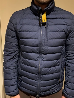 Mörkblå dunjacka Parajumpers M - Mörkblå dunjacka från Parajumpers i storlek M. Jackan är quiltad med horisontella sömmar, har hög krage med gul detalj och dragkedja framtill. Märket på ärmen