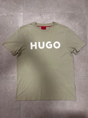 Grön HUGO t-shirt med tryck - Snygg grön t-shirt från HUGO med stort vitt logotryck på bröstet. Klassisk rund hals och korta ärmar. Tillverkad i mjuk bomull som känns skön mot huden. Perfekt för dig som gillar stilrena och enkla plagg med tydlig branding.