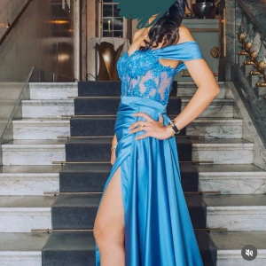 Blå långklänning med spets och pärlor - Säljer en elegant blå långklänning med offshoulder och slits. Överdelen har transparent spets med blommiga broderier och pärlor, samt en vacker omlott-effekt i midjan. Kjolen är gjord i glansigt satinmaterial och faller snyggt. Perfekt för bal eller fest.
