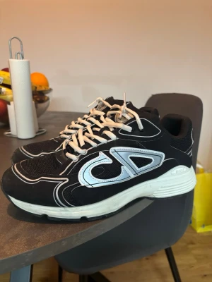 B30 svarta sneakers med vita detaljer - Säljer ett par B30 sneakers i svart med vita och silverfärgade detaljer. Skorna har en chunky vit sula, meshpartier och tydliga CD-loggor på sidorna och hälen. Klassisk snörning och sportig design som sticker ut.