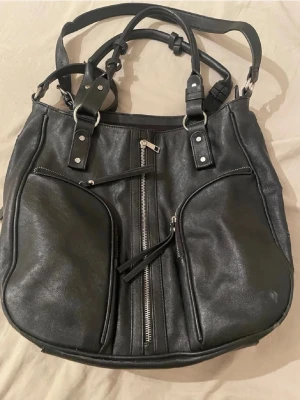 Svart axelväska med dragkedjor - Säljer en svart axelväska i skinnimitation med flera snygga dragkedjor och detaljer i silver. Väskan har två stora ytterfack med dragkedja, innerfack och rejäla handtag med metallringar. Perfekt för dig som vill ha plats för allt och gillar en cool stil. Lite sliten på kanten uppe även handtagen se bilderna. Köpt sacond hand