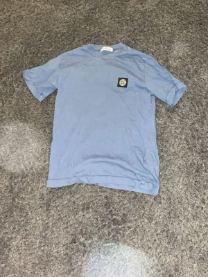 Blå t-shirt från Stone Island - Säljer en blå t-shirt från Stone Island med klassisk logopatch på bröstet. T-shirten har korta ärmar och en rund hals. Perfekt för dig som gillar streetwear och vill ha något stilrent med ikonisk branding. Bra skick. Använd några gånger. Storlek 13-15 år. 200kr