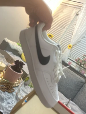 Nike Court Vision Low sneakers vita - Nike Court Vision Low sneakers i vit färg med svart swoosh på sidan. Klassisk låg modell med snörning och perforerade detaljer på tån för extra ventilation. Platt sula och rund tå. Tillverkade i syntetmaterial och skinn. Kommer i originalkartong.