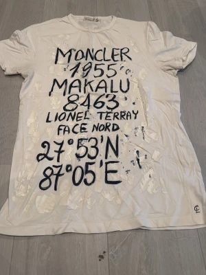Vit Moncler t-shirt - Säljer en vit t-shirt från Moncler i storlek M med coolt handmålat tryck och text framtill. T-shirten har korta ärmar och rund hals. Materialet är mjuk bomull och designen har unika vita och svarta detaljer som ger en konstnärlig vibe. Köpt i Italien på semester. Ny pris 3800kr, limited edition.