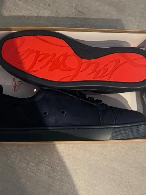 Mörkblå sneakers från Christian Louboutin - Snygga mörkblå sneakers från Christian Louboutin med klassisk röd sula och broderad logga på hälen. Skorna är gjorda i mocka och har rund tå, snörning och platt sula. Insidan är beige och har exklusiv känsla. Perfekt för dig som gillar lyxiga detaljer.